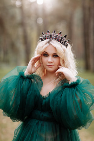 A beautiful blonde young woman in a long green dress and a diadem on her head in the forest. girl sitting near the old stump. Solar glare. Fantasy. fairy tale.の写真素材