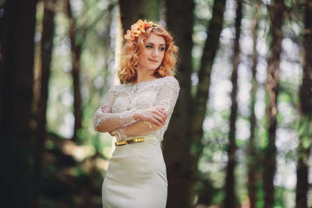 beautiful young red-haired bride in the forest with a floral wreath on her head. woman in long white dress outdoors on summer day. wedding day.の写真素材