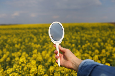 A female hand with a miniature retro mirror in field of yellow wildflowers is reflected. Creative...