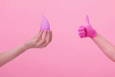 Hands of a young woman in pink latex gloves and hand with menstrual cup on pink background. women's health. preservation of ecology. waste-free use. women's hygiene products.の写真素材