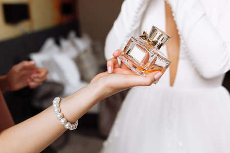 A bridesmaid's hand holding a bottle with perfume on bride in white dress background. Morning of the bride in the house, hands without face. wedding day. mock up spaceの写真素材