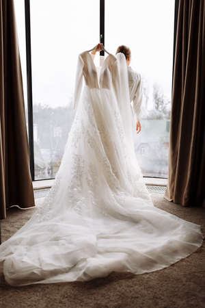 Morning of the bride. the bride is holding a luxury white long dress in her hands by the large window in hotel room. back viewの写真素材