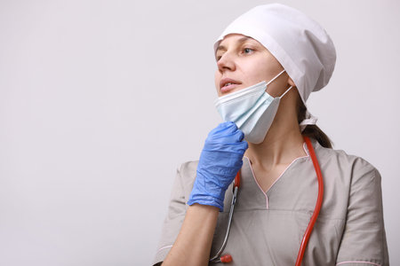 smiling female doctor with stethoscope in uniform. Woman is taking off medical protective mask from face to breathe air, on white background. Difficulties and problems at work of a medical workerの写真素材