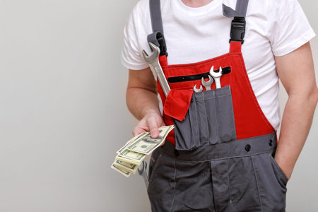 cropped photo of worker in red uniform, protective hard hat holding bundle of dollars, cash money on...