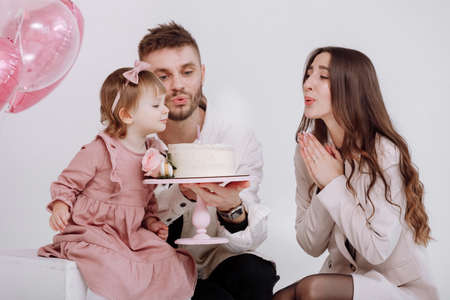 young parents mom and dad are celebrating birthday their two years old child. family light and blowing out the candles on the cake with flowers on the background of birthday decor with pink ballsの写真素材