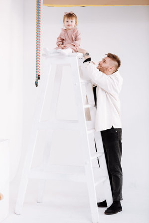 stylish man is having fun with cute child baby girl in decorated studio. daughter tries to climb the...