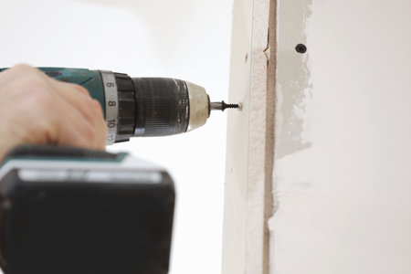 The installer worker in safe medical mask and overalls fixes a detail of the stiffener element with screws using an electric screwdriver to the wall of the metal structure.の写真素材