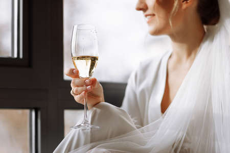 Bride s morning. Bride drinking champagne in the peignoir. young woman is sitting on a large window in a hotel room in bathroom. Beautiful girl in white wedding robe. wedding day.の写真素材