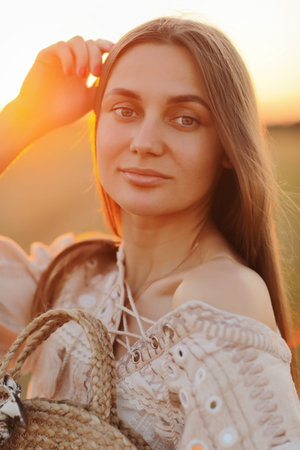 beautiful young woman with long hair and straw bag in hand in the summer at sunset in the field for...