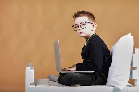 schoolboy in glasses using laptop sitting on bed. Child doing school homework and seriously and...