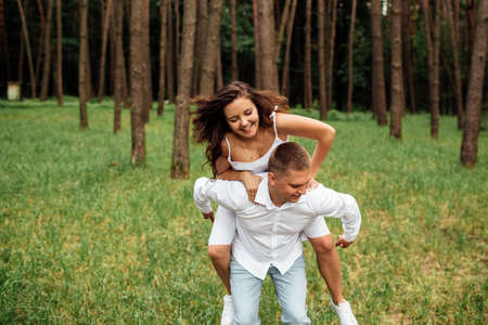 Smiling couple having fun in the park. Love and tenderness, dating, romance, lovers enjoy each other in the summer park. man carry on his back the beautiful curly hair woman smiling and laughingの写真素材