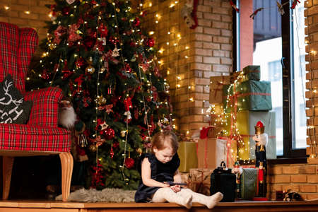 Merry Christmas celebration. Beautiful little girl in a dress sitting near the Christmas tree. Happy little smiling girl using cellphone. Christmas miracles. Happy New Yearの写真素材