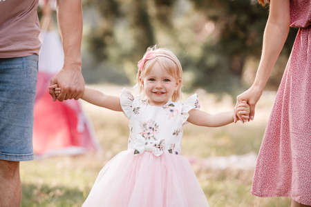 View on toddler. Mother, father hold hands daughter enjoy nature and walk in the park. Young family spending time together on vacation, outdoors. The concept of family summer holidayの写真素材