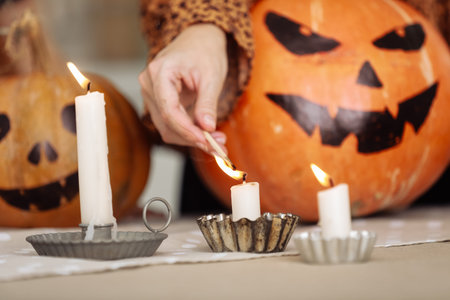 holidays and leisure concept - woman's hand with matches lighting candles at home on halloween. painted pumpkins on a background. horror theme and Hallowe'enの写真素材
