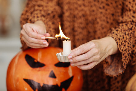 holidays and leisure concept - woman's hand with matches lighting candles at home on halloween. painted pumpkins on a background. horror theme and Hallowe'enの写真素材