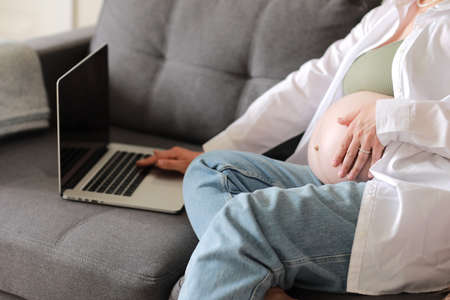 Attractive pregnant young lady working remotely online from home. Close up woman hands on laptop with big belly advanced pregnancyの写真素材