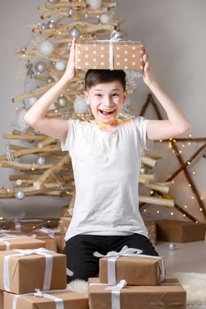 Holidays Concept. A smiling excited boy child by the wooden decoration Christmas tree, in his hands he holds a big gift box. new Year's Eve and Christmas, waiting for a miracle.の写真素材