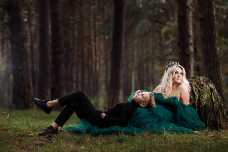 A beautiful blonde young woman in a long green dress and a diadem on her head with stylish young son in the forest. girl and boy sitting near the old stump. solar glare. Fantasy. fairy tale.の写真素材