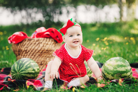 Cute 10 months baby girl on picnic on grass with watermelons. child in red dress and bow on head. Concept of healthy eating, childhood, fruit ripeness, parenting, seasonal harvestの写真素材