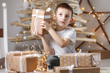 Holidays Concept. A smiling little excited boy child is sitting by the wooden decorations Christmas tree and many gift boxes, holding a gift box. new Year's Eve and Christmas, waiting for a miracle.の写真素材