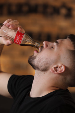 RIVNE, UKRAINE JUNE 26 2020: young bearded man drinking coca cola from bottle in barbershop on brick wall background.のeditorial素材