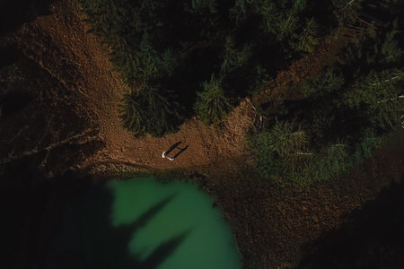 Aerial view of two people standing by the natural lake surrounded by pine trees in Karpaty...