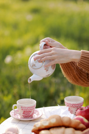 female hands pour tea into a cup from a teapot. Breakfast picnic with croissants and tea in spring blooming park on a white tablecloth on a sunny day. Outdoor, picnic, brunch, spring mood.の写真素材