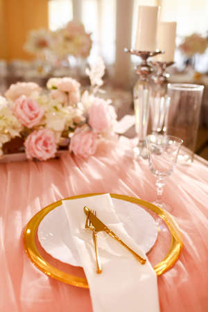 table decoration with pink tablecloth, crystal candlesticks with candles and white pink flowers in restaurant . stylish wedding day. setting table with gold plate, fork and knife.の写真素材