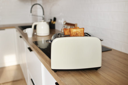 Modern white toaster and roasted bread slices toasts inside on wooden table in kitchen.