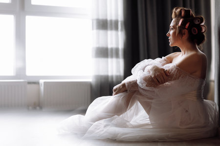 A beautiful young showy brunette woman with bright pin-up make-up in long white lace dress and pink curlers. beauty concept. girl isolated on gray background. international women's dayの写真素材