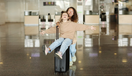 Summer Vacation Concept. Portrait of cheerful positive teen girl having fun and spreading hands, ready for a flight, sitting on suitcase. mom is pushing baggage, riding daughter.の写真素材