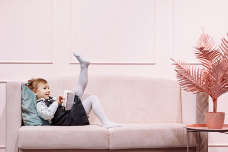Little cute adorable girl is lying alone on pink sofa and reading book for childs in living room with decorの写真素材