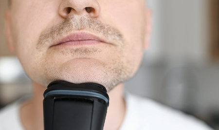 morning routine hygiene. young handsome man in white t shirt is shaving beard with electric razor...