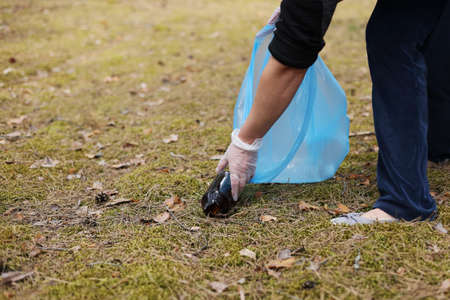 A woman hands in gloves collects and puts used plastic bottle into a blue trash bag. A volunteer cleans up the park on a sunny bright day. Clearing, pollution, ecology and plastic concept.の写真素材