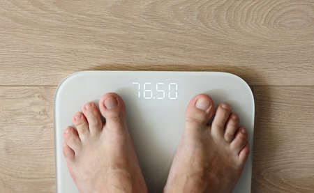 Man weighing himself - male bare feet stepping on white digital floor scales at home: close up view. Measuring weight, control, wellness and diet concept.の写真素材