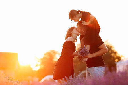 happy family day. Young mother and father carrying on shoulders daughter in lavender field on sunset. Dad, mom and child girl kissing and hugging on nature on summer. friendly family. family look.の写真素材