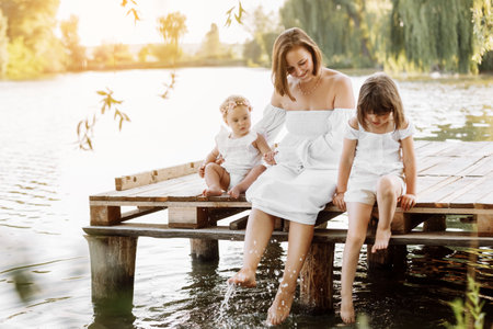 mother day. Happy young mom with little girls sitting on wooden bridge near lake, pond on summer. Family enjoying life together. Mother and two child daughter smiling while spending free time outdoorsの写真素材