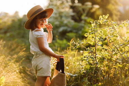 Portrait of cute little girl is smelling wild flower and walks in the rays of a sunset in a meadow flowering, enjoying the summer, warmth, freedom. Child in mommys big straw hat having fun outdoors.の写真素材