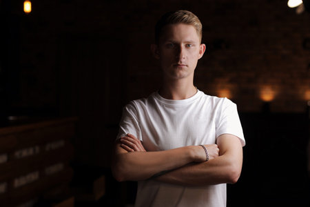 portrait of serious young man with folded and crossing hands standing on dark background. Guy in...