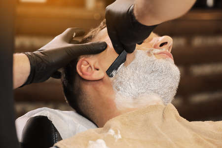 Barber Man is shaving client with a vintage straight razor in a barbershop. Shaving cream, foam. Classic shave by stainless steel straight edge razorの写真素材