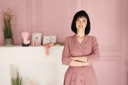 Portrait of attractive smiling brunette with short hair woman folded arms staying alone at stylish interior indoors. Female in soft pink dress is looking in camera.の写真素材