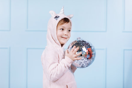 Smiling beautiful little girl in unicorn pink costume is smiling and having fun with disco ball on a blue wall background. happy childhoodの写真素材