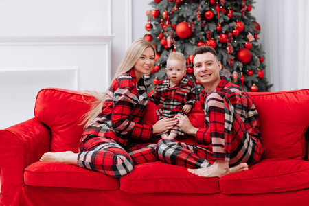 Young happy family in the same pajamas: mom, dad, baby boy are lying down on the floor by the festive Christmas tree and red sofa. Family holiday is New year and Christmasの写真素材