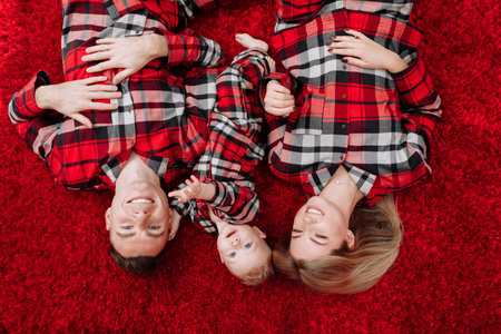 Young happy family in the same pajamas: mom, dad, baby boy are lying down on the floor by the festive Christmas tree and red sofa. Family holiday is New year and Christmasの写真素材