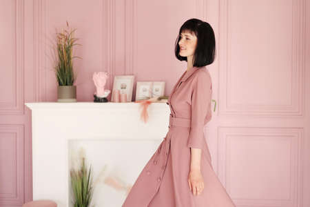 Portrait of attractive smiling brunette with short hair woman folded arms staying alone at stylish interior indoors. Female in soft pink dress is looking in camera.の写真素材