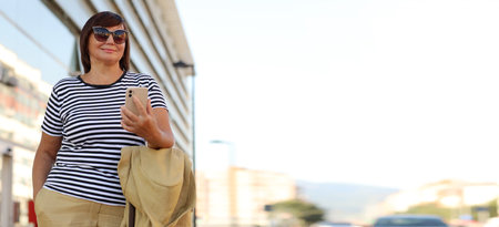 Ready for business trip. mature stylish business woman in sunglasses in red suitcase luggage. smiling female waiting for taxi and uses smartphone outdoors by the airport or railway station.の写真素材