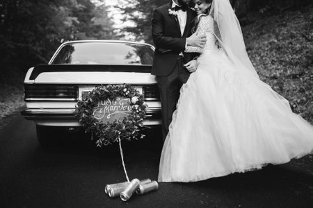 black and white photo of happy young newlywed couple near decorated retro car with plate just married and many jars, outdoors. Bride in white long dress and groom in suit are hugging in Wedding dayの写真素材