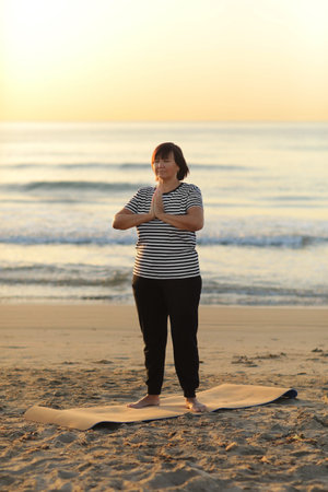 Yoga and meditation of mature woman practicing breath exercises on the sea beach on sunrise in the...