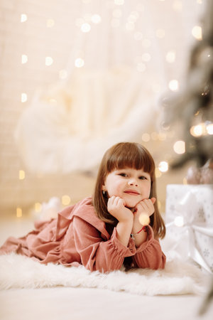 little cute child girl in pink dress lying on white furry carpet in decorated Christmas room interior and looking in camera. beautiful bokeh. Happy family New Year, Xmas concept.の写真素材
