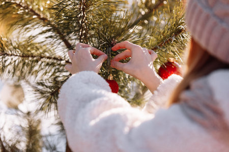 Child girl is decorating Christmas tree with shiny red balls outdoors before the holidays. Kid in warm jacket and hat is having fun outside in winter day. Merry Christmas and happy family holidaysの写真素材